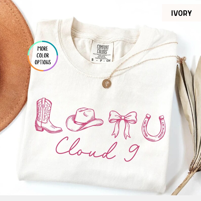 May include: Ivory-colored t-shirt with a pink graphic of a cowboy boot, hat, bow, and horseshoe, with the words "Cloud 9" in cursive. The shirt has a crew neck and short sleeves. A gold necklace is draped on the shirt.