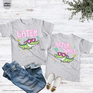 May include: Two athletic heather gray t-shirts with a cartoon alligator wearing pink sunglasses. One shirt says "LATER" and the other says "AFTER WHILE". The shirts are paired with blue jeans and white sneakers.