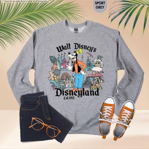 Pu&ograve; includere: Una felpa grigia con una grafica di Pippo davanti alle attrazioni di Disneyland. Il testo "Walt Disney's Disneyland Est.1955" &egrave; stampato sotto. La felpa &egrave; abbinata a jeans blu scuro, occhiali con montatura arancione e scarpe da ginnastica arancioni.
