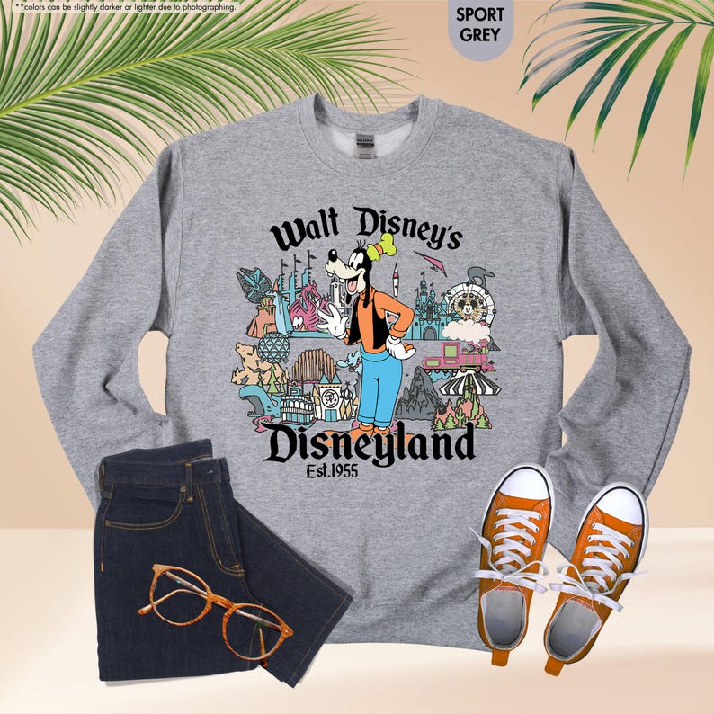 Pu&ograve; includere: Una felpa grigia con una grafica di Pippo davanti alle attrazioni di Disneyland. Il testo "Walt Disney's Disneyland Est.1955" &egrave; stampato sotto. La felpa &egrave; abbinata a jeans blu scuro, occhiali con montatura arancione e scarpe da ginnastica arancioni.
