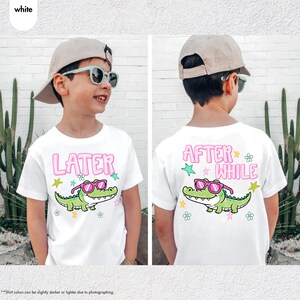 May include: White t-shirts with a cartoon alligator wearing sunglasses. The front of the shirt says "LATER" in pink letters, and the back says "AFTER WHILE". The alligator is green with pink sunglasses.
