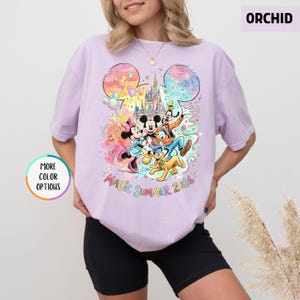 K&ouml;nnte beinhalten: Orchideenfarbenes T-Shirt mit einem farbenfrohen Aufdruck von Mickey Mouse, Minnie Mouse, Goofy, Donald Duck und Pluto vor einem Schloss. Die Worte "Magic Summer 2026" sind darunter gedruckt. Das T-Shirt hat kurze &Auml;rmel.