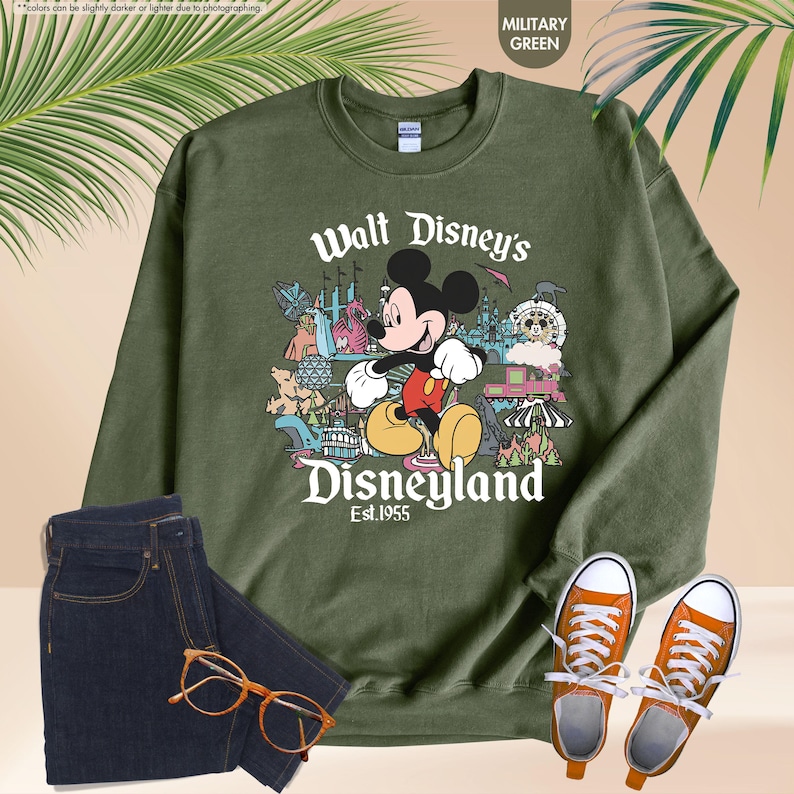 Pu&ograve; includere: Felpa verde militare con Topolino e la scritta "Walt Disney's Disneyland Est.1955". L'immagine mostra anche jeans blu scuro, scarpe da ginnastica arancioni e occhiali. Sullo sfondo &egrave; visibile una foglia di palma.