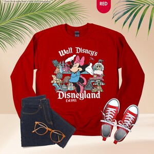 Pu&ograve; includere: Una felpa rossa con una grafica di Minnie Mouse e la scritta "Walt Disney's Disneyland Est. 1955". L'immagine include anche un paio di jeans blu, scarpe da ginnastica rosse e occhiali con montatura dorata.