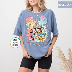 K&ouml;nnte beinhalten: Blaues Jeans-T-Shirt mit einer farbenfrohen Grafik von Mickey Mouse, Minnie Mouse, Donald Duck, Goofy und Pluto vor einem Schloss. Der Text "MAGIC SUMMER 2026" befindet sich unter den Figuren. Das Shirt hat kurze &Auml;rmel.