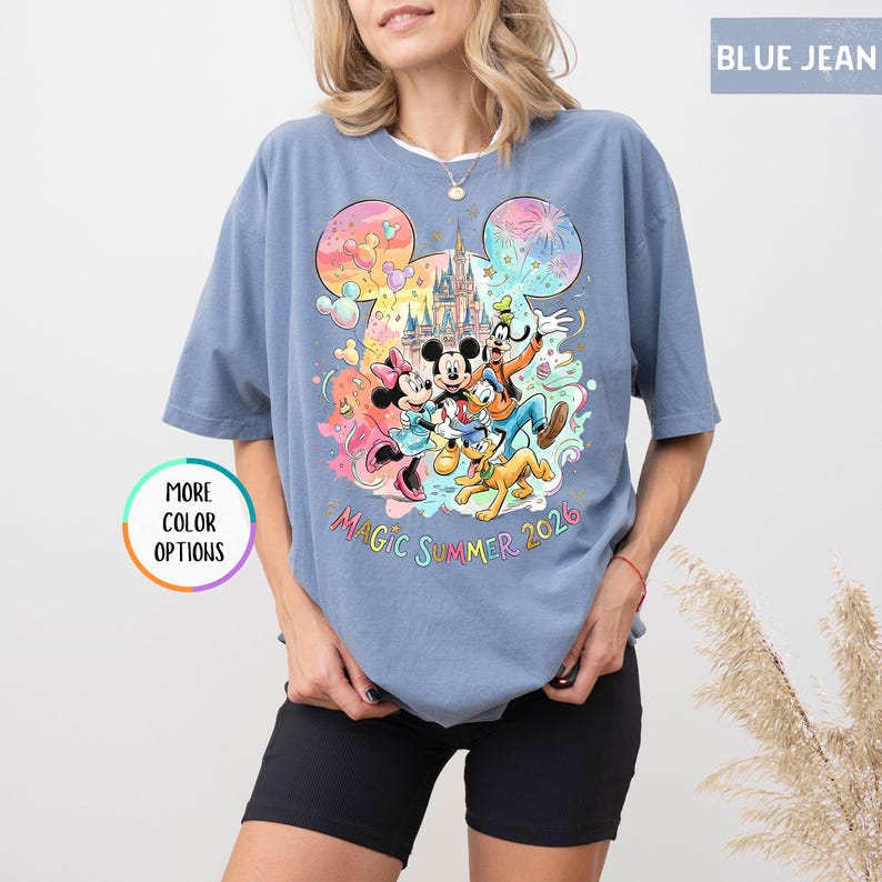 K&ouml;nnte beinhalten: Blaues Jeans-T-Shirt mit einer farbenfrohen Grafik von Mickey Mouse, Minnie Mouse, Donald Duck, Goofy und Pluto vor einem Schloss. Der Text "MAGIC SUMMER 2026" befindet sich unter den Figuren. Das Shirt hat kurze &Auml;rmel.