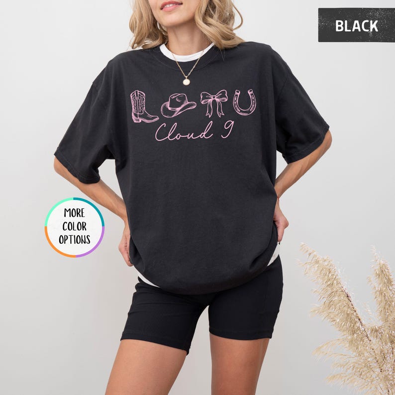 May include: Black t-shirt with a white collar featuring a pink graphic design of a cowboy boot, hat, bow, and horseshoe, with the text "Cloud 9". The shirt is a relaxed fit.