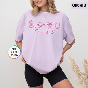 May include: A light purple t-shirt with a pink graphic of a cowboy boot, hat, bow, and horseshoe, with the words "Cloud 9" in a cursive font. The shirt is a solid color and has short sleeves.