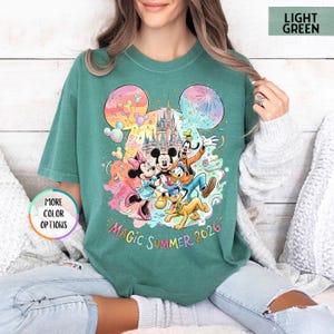 K&ouml;nnte beinhalten: Hellgr&uuml;nes T-Shirt mit einem farbenfrohen Disney-Motiv, das Micky Maus, Minnie Maus, Donald Duck, Goofy und Pluto zeigt. Das Design beinhaltet ein Schloss, Feuerwerk und den Text "Magic Summer 2026".