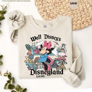 Pu&ograve; includere: Una felpa color sabbia con una grafica di Minnie Mouse con un vestito blu e un fiocco rosa, con la scritta "Walt Disney's Disneyland Est.1955". Il design include varie icone di Disneyland.