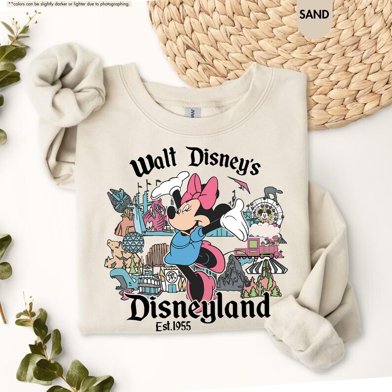 Pu&ograve; includere: Una felpa color sabbia con una grafica di Minnie Mouse con un vestito blu e un fiocco rosa, con la scritta "Walt Disney's Disneyland Est.1955". Il design include varie icone di Disneyland.