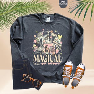 以下が含まれることがあります： 「The Most Magical Place」と書かれたカラフルなグラフィックが特徴の濃いヘザーグレーのスウェットシャツ。デザインにはお城、ミッキーマウスの耳、その他のテーマパークの要素が含まれています。画像には、濃い色のジーンズ、オレンジ色のスニーカー、メガネも写っています。