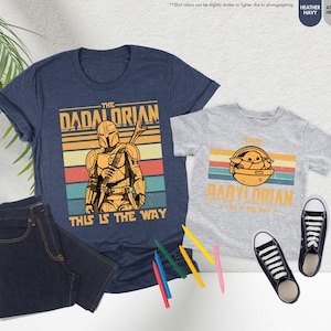 以下が含まれることがあります： 「The Dadalorian」と「The Babylorian」のグラフィックがプリントされた、ネイビーブルーとヘザーグレーのTシャツ。濃い色のジーンズ、白と黒のスニーカー、色鉛筆も画像に写っています。