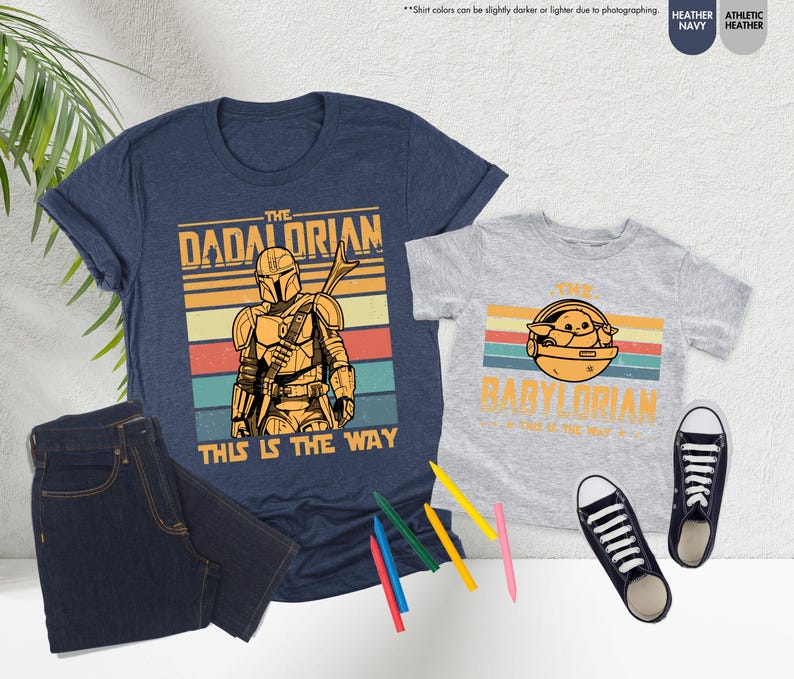 以下が含まれることがあります： 「The Dadalorian」と「The Babylorian」のグラフィックがプリントされた、ネイビーブルーとヘザーグレーのTシャツ。濃い色のジーンズ、白と黒のスニーカー、色鉛筆も画像に写っています。