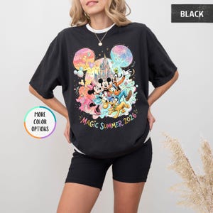 K&ouml;nnte beinhalten: Schwarzes T-Shirt mit einem farbenfrohen Aufdruck von Mickey Mouse, Minnie Mouse, Donald Duck, Goofy und Pluto vor einem Schloss. Der Text "Magic Summer 2026" ist darunter gedruckt. Das T-Shirt hat einen Rundhalsausschnitt und kurze &Auml;rmel.