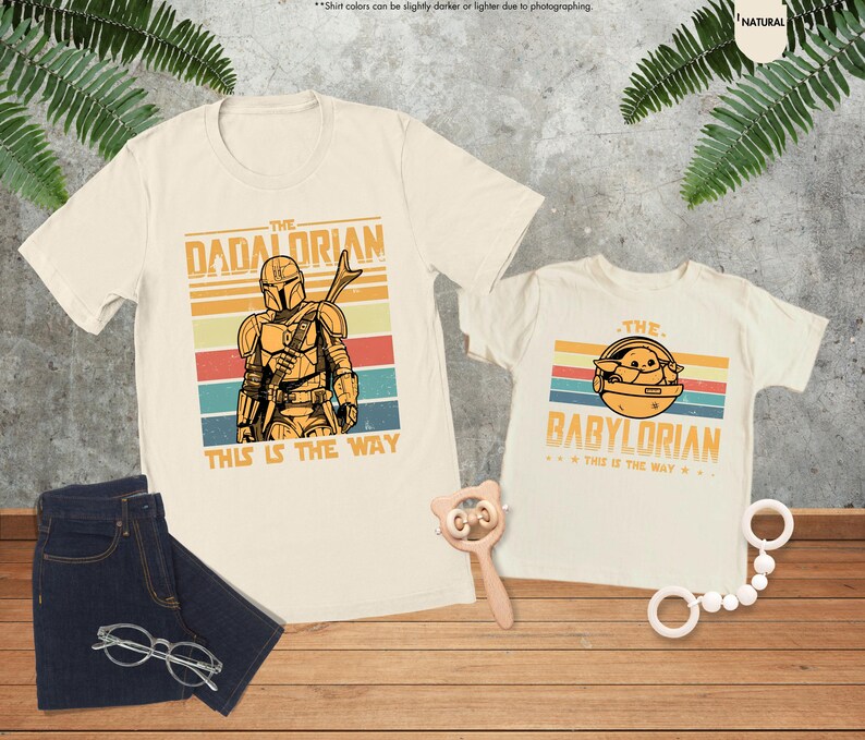 以下が含まれることがあります： スターウォーズをテーマにしたグラフィックが施された、クリーム色のTシャツ2枚。「The Dadalorian」とマンダロリアンの姿が描かれたシャツと、「The Babylorian」とグローグーのイメージが描かれたシャツ。どちらのシャツにも「This is the Way」というフレーズが書かれています。