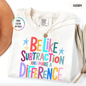 May include: An ivory t-shirt featuring the multi-colored text BE LIKE SUBTRACTION AND MAKE A DIFFERENCE. The shirt has a Comfort Colors brand label at the neck and includes small blue and yellow star graphics. The design uses various bright colors.