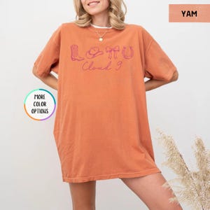 May include: An orange-colored t-shirt with a graphic design in purple. The design includes a cowboy boot, a hat, a bow, and a horseshoe, with the words "Cloud 9" in cursive. The shirt has short sleeves and a relaxed fit.