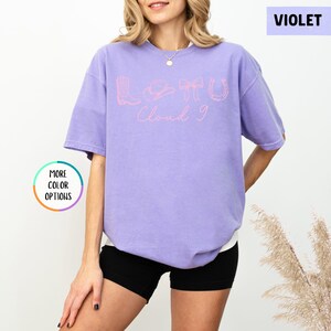 May include: A violet short-sleeved t-shirt with the text "Lotto Cloud 9" in pink. The design includes a boot, a cowboy hat, and a horseshoe. The shirt is a relaxed fit and has a round neckline.
