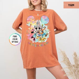 K&ouml;nnte beinhalten: Ein orangefarbenes T-Shirt mit einem farbenfrohen Aufdruck von Disney-Figuren, darunter Micky und Minnie Maus, Goofy und Pluto, vor einem Schloss. Der Text "Magic Summer 2026" ist unter den Figuren aufgedruckt.
