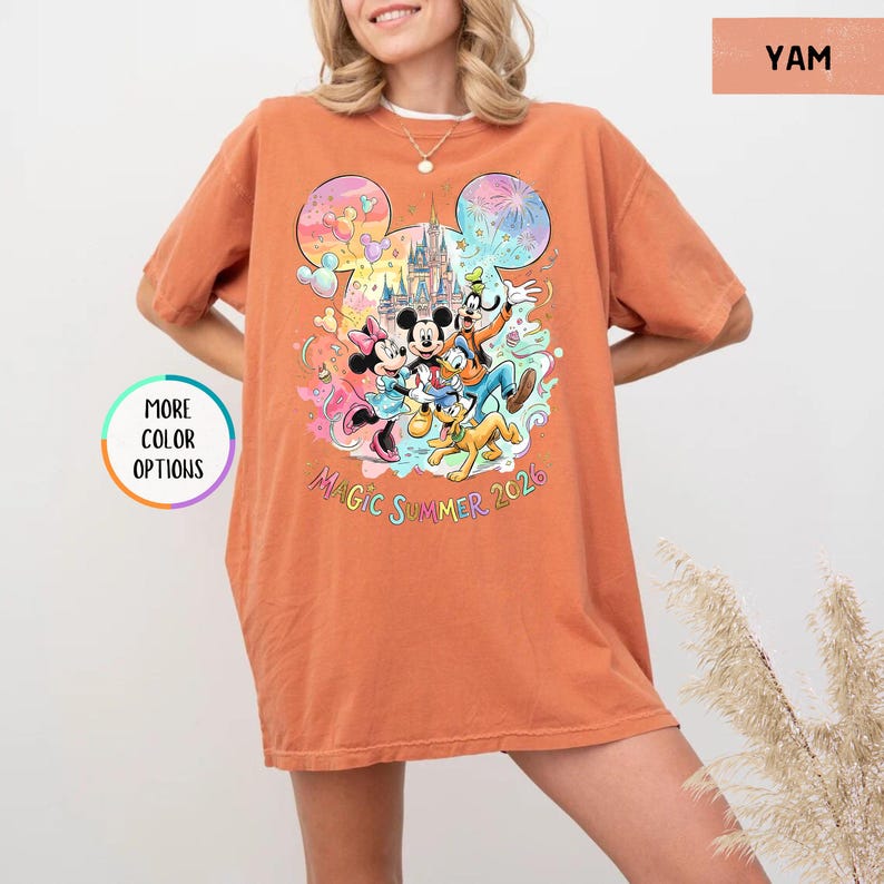 K&ouml;nnte beinhalten: Ein orangefarbenes T-Shirt mit einem farbenfrohen Aufdruck von Disney-Figuren, darunter Micky und Minnie Maus, Goofy und Pluto, vor einem Schloss. Der Text "Magic Summer 2026" ist unter den Figuren aufgedruckt.