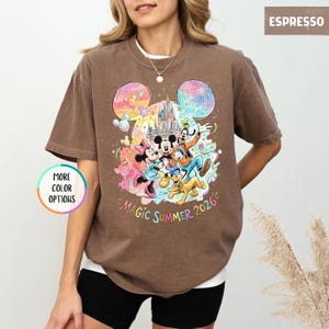 K&ouml;nnte beinhalten: Espressofarbenes T-Shirt mit einem farbenfrohen Aufdruck von Disney-Figuren, darunter Mickey und Minnie Mouse, Goofy und Donald Duck. Das Design beinhaltet ein Schloss und den Text "Magic Summer 2026". Das Shirt hat kurze &Auml;rmel und einen Rundhalsausschnitt.