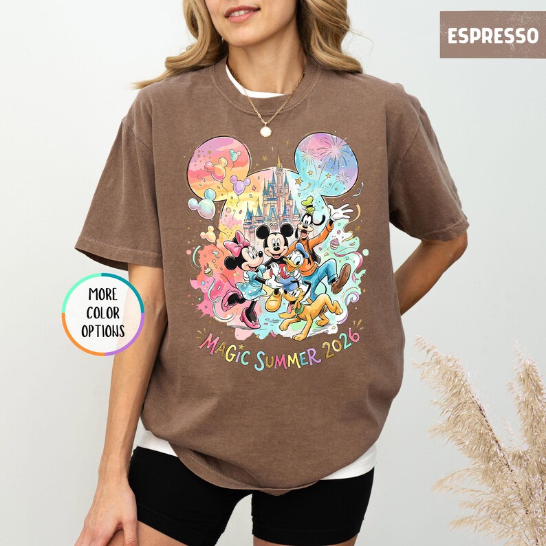 K&ouml;nnte beinhalten: Espressofarbenes T-Shirt mit einem farbenfrohen Aufdruck von Disney-Figuren, darunter Mickey und Minnie Mouse, Goofy und Donald Duck. Das Design beinhaltet ein Schloss und den Text "Magic Summer 2026". Das Shirt hat kurze &Auml;rmel und einen Rundhalsausschnitt.