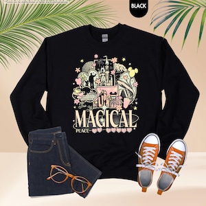 以下が含まれることがあります： お城、ミッキーマウス、そして「The Most Magical Place」の文字が描かれた、カラフルなグラフィックデザインの黒い長袖スウェットシャツ。スウェットシャツは、青いジーンズ、オレンジ色のメガネ、オレンジ色のスニーカーと組み合わせています。
