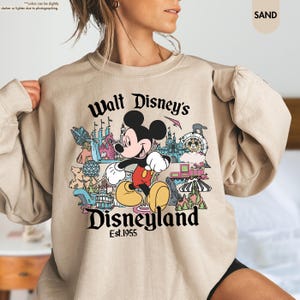 Pu&ograve; includere: Felpa color sabbia con una grafica colorata di Topolino davanti alle attrazioni di Disneyland. Il testo "Walt Disney's Disneyland Est.1955" &egrave; stampato sotto. La felpa ha un girocollo e maniche lunghe.