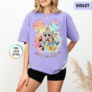 K&ouml;nnte beinhalten: Ein violettes T-Shirt mit einem farbenfrohen Aufdruck von Disney-Figuren, darunter Micky und Minnie Maus, Donald Duck und Goofy, vor einem Schloss. Der Text "MAGIC SUMMER 2026" ist darunter. Das Shirt hat eine lockere Passform.