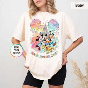 K&ouml;nnte beinhalten: Elfenbeinfarbenes T-Shirt mit einer farbenfrohen Grafik von Mickey Mouse, Minnie Mouse, Donald Duck, Goofy und Pluto vor einem Schloss. Der Text "Magic Summer 2026" steht darunter. Das Shirt hat kurze &Auml;rmel.