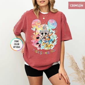 K&ouml;nnte beinhalten: Karmesinrotes T-Shirt mit einem farbenfrohen Aufdruck von Mickey Mouse, Minnie Mouse, Donald Duck, Goofy und Pluto vor einem Schloss. Der Text "Magic Summer 2026" steht darunter. Das T-Shirt hat kurze &Auml;rmel.