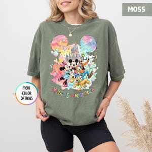 K&ouml;nnte beinhalten: Moosgr&uuml;nes T-Shirt mit einem farbenfrohen Aufdruck von Mickey Mouse, Minnie Mouse, Donald Duck, Goofy und Pluto vor einem Schloss. Der Text "MAGIC SUMMER 2026" steht darunter. Das Shirt hat kurze &Auml;rmel.