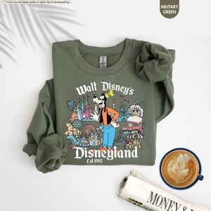 Pu&ograve; includere: Felpa verde militare con una grafica colorata di Pippo e delle attrazioni di Disneyland. Il testo "Walt Disney's Disneyland Est. 1955" &egrave; stampato sul davanti. Una tazza di caff&egrave; e un giornale sono nell'immagine.