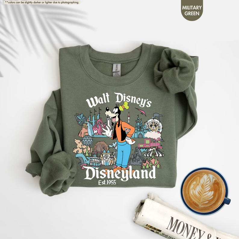 Pu&ograve; includere: Felpa verde militare con una grafica colorata di Pippo e delle attrazioni di Disneyland. Il testo "Walt Disney's Disneyland Est. 1955" &egrave; stampato sul davanti. Una tazza di caff&egrave; e un giornale sono nell'immagine.