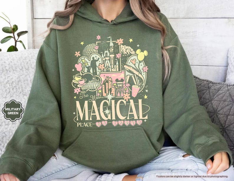 以下が含まれることがあります： ミリタリーグリーンのパーカー。お城、ミッキーマウス、そして「The Most Magical Place」の文字が描かれたグラフィックデザイン。ピンク、イエロー、ホワイトの要素が含まれています。パーカーにはフロントポケットとドローストリングフードが付いています。