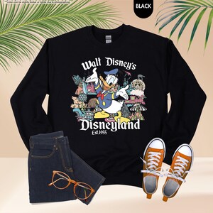 Pu&ograve; includere: Felpa nera con grafica di Paperino davanti a Disneyland. Il testo "Walt Disney's Disneyland Est.1955" &egrave; stampato sotto. L'immagine include anche jeans blu scuro, occhiali arancioni e scarpe da ginnastica arancioni.