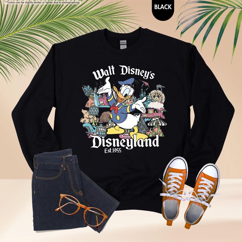 Pu&ograve; includere: Felpa nera con grafica di Paperino davanti a Disneyland. Il testo "Walt Disney's Disneyland Est.1955" &egrave; stampato sotto. L'immagine include anche jeans blu scuro, occhiali arancioni e scarpe da ginnastica arancioni.