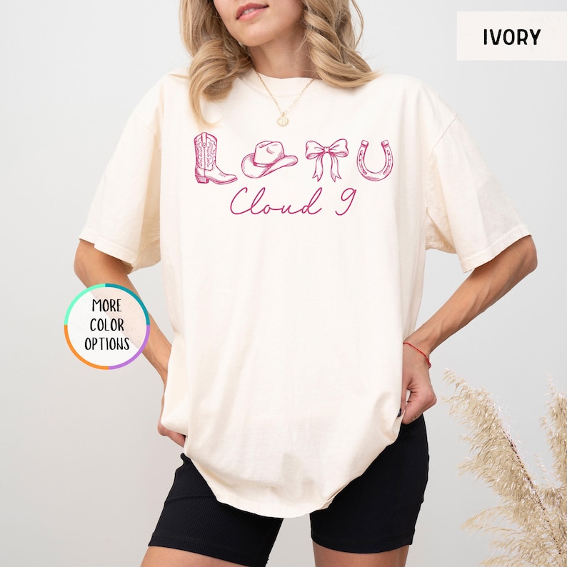 May include: Ivory-colored t-shirt with a pink graphic of a cowboy boot, hat, bow, and horseshoe, with the words "Cloud 9" in cursive. The shirt has short sleeves and a relaxed fit.