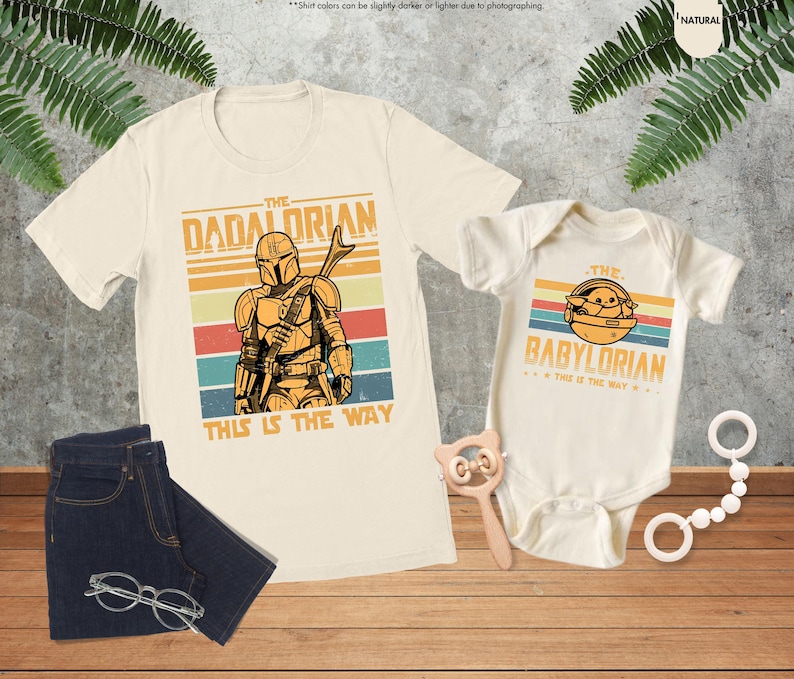 以下が含まれることがあります： スターウォーズをテーマにしたグラフィックが特徴の、クリーム色のTシャツとベビーボディスーツのセットです。大人用Tシャツには「The Dadalorian」、ベビーボディスーツには「The Babylorian」と書かれています。青いジーンズとメガネも画像にあります。
