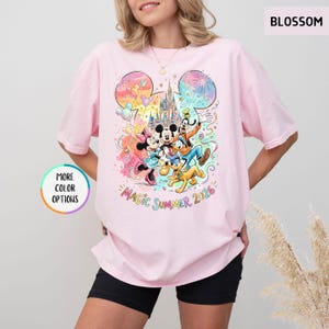 K&ouml;nnte beinhalten: Ein hellrosa T-Shirt mit einem farbenfrohen Aufdruck von Disney-Figuren, darunter Micky und Minnie Maus, Goofy, Donald Duck und Pluto. Das Design zeigt ein Schloss, Feuerwerk und den Text "Magic Summer 2026".