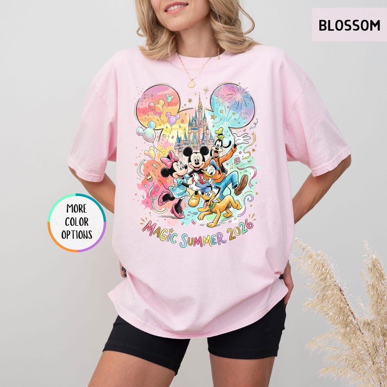 K&ouml;nnte beinhalten: Ein hellrosa T-Shirt mit einem farbenfrohen Aufdruck von Disney-Figuren, darunter Micky und Minnie Maus, Goofy, Donald Duck und Pluto. Das Design zeigt ein Schloss, Feuerwerk und den Text "Magic Summer 2026".