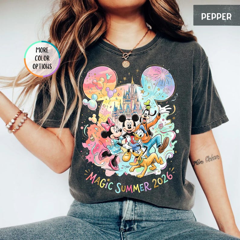 K&ouml;nnte beinhalten: Dunkelgraues T-Shirt mit einem farbenfrohen Aufdruck von Mickey Mouse, Minnie Mouse, Donald Duck, Goofy und Pluto vor einem Schloss. Der Text "Magic Summer 2026" ist darunter gedruckt. "More Color Options" steht in einem Kreis links.