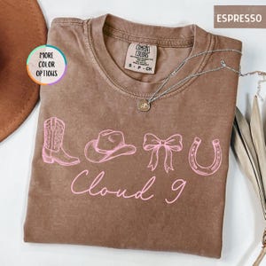 May include: Espresso-colored t-shirt with a pink graphic of a cowboy boot, hat, bow, and horseshoe. The words "Cloud 9" are written in pink script below the graphic. A silver necklace with a pendant is visible.