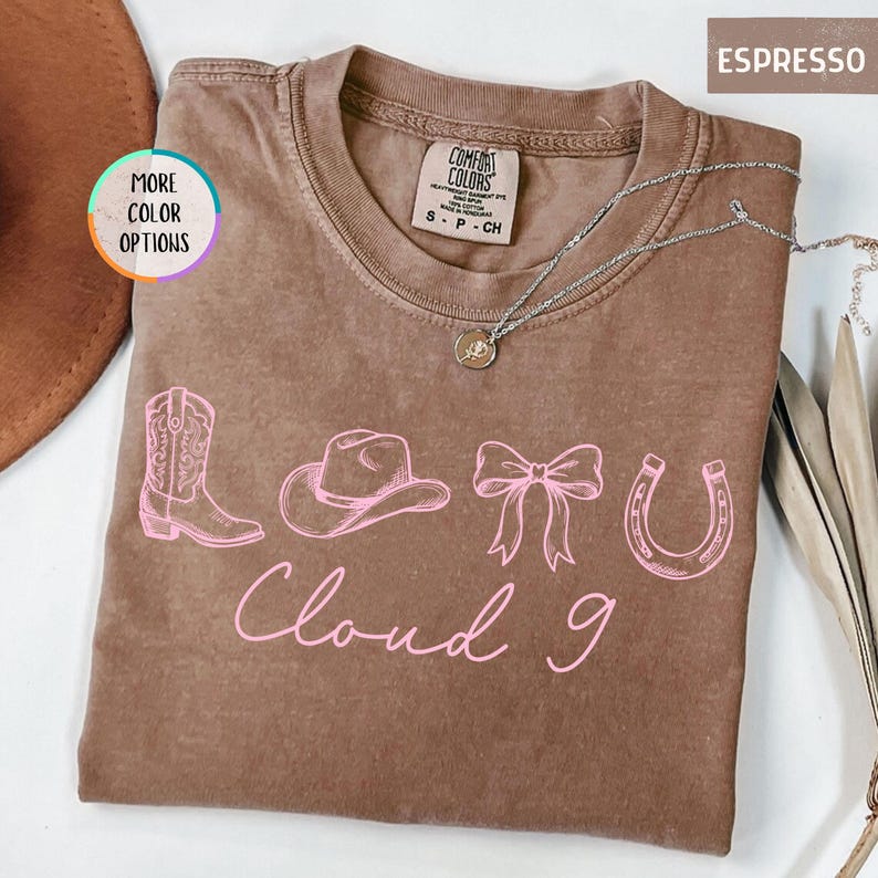 May include: Espresso-colored t-shirt with a pink graphic of a cowboy boot, hat, bow, and horseshoe. The words "Cloud 9" are written in pink script below the graphic. A silver necklace with a pendant is visible.