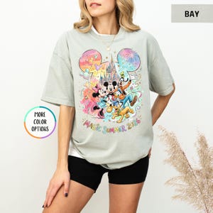 K&ouml;nnte beinhalten: Hellgraues T-Shirt mit einem farbenfrohen Aufdruck von Mickey Mouse, Minnie Mouse, Goofy, Pluto und einem Schloss. Die Worte "Magic Summer 2026" sind darunter gedruckt. Das T-Shirt hat kurze &Auml;rmel und einen Rundhalsausschnitt.