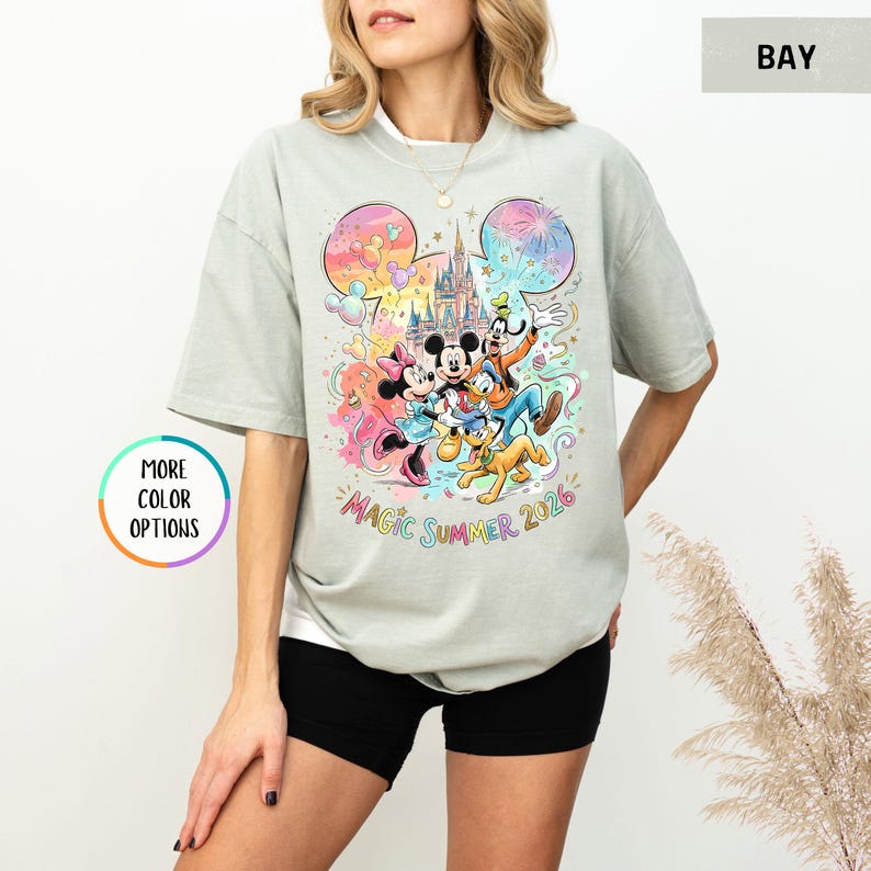 K&ouml;nnte beinhalten: Hellgraues T-Shirt mit einem farbenfrohen Aufdruck von Mickey Mouse, Minnie Mouse, Goofy, Pluto und einem Schloss. Die Worte "Magic Summer 2026" sind darunter gedruckt. Das T-Shirt hat kurze &Auml;rmel und einen Rundhalsausschnitt.