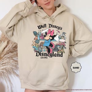 Pu&ograve; includere: Felpa con cappuccio color sabbia con una grafica di Minnie Mouse davanti a Disneyland. Il testo "Walt Disney's Disneyland Est. 1955" &egrave; stampato sopra e sotto l'immagine. La felpa ha una tasca a marsupio e un cappuccio con coulisse.
