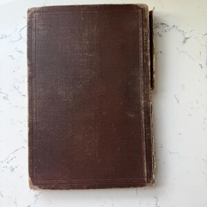 May include: A vintage, rectangular book with a worn, dark brown cover. The cover has a textured appearance with visible wear and tear, suggesting age. The edges of the book show signs of use, adding to its antique aesthetic. The book is likely a collectible or decorative item.