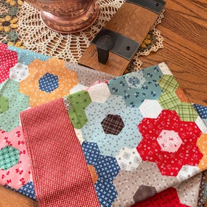 May include: A fabric quilt featuring a patchwork of multicolored hexagonal shapes in floral, plaid, and geometric patterns. The quilt has a prominent red and white gingham border and is displayed alongside a wooden board with a metal hook.